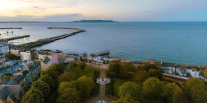 An elevated coastal panorama captures a manicured green park centered around a fountain, bordered by seaside streets and a long stone pier extending into the deep blue ocean.