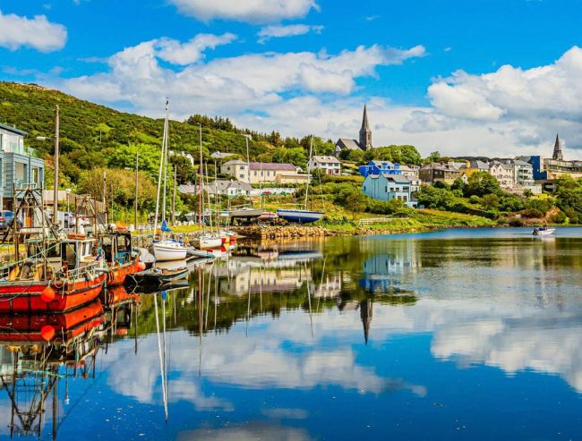 A picturesque waterfront in a coastal Irish town features colorful boats gently moored in a tranquil bay, while vibrant buildings and a church spire are mirrored beautifully in the glass-like water.