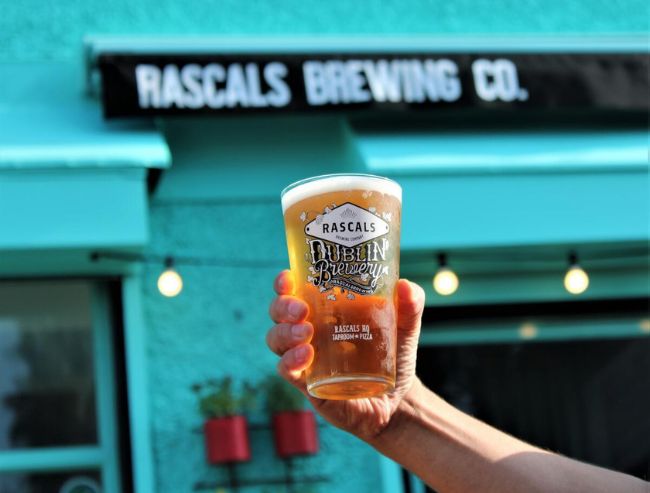 A hand holding up a full glass of beer featuring a "Rascals Dublin Brewery" logo against a bright teal wall.