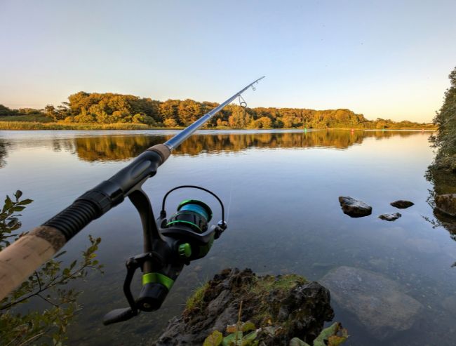 Point-of-view shot of a spinning fishing rod and reel extending over a calm river reflecting autumn-colored trees on the far bank.