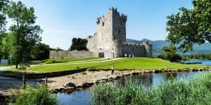 A historic stone castle stands on a grassy hill beside a calm lake, surrounded by vibrant green trees and distant mountains under a bright, clear blue sky.