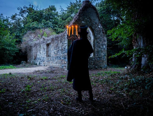 A silhouetted figure in a top hat and long coat stands in a dark forest, holding a lit candelabra before old, overgrown stone ruins.